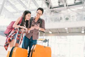 Asian couple travelers using smartphone checking flight or online check-in at airport, with passport and luggage. Air travel or mobile phone technology concept.